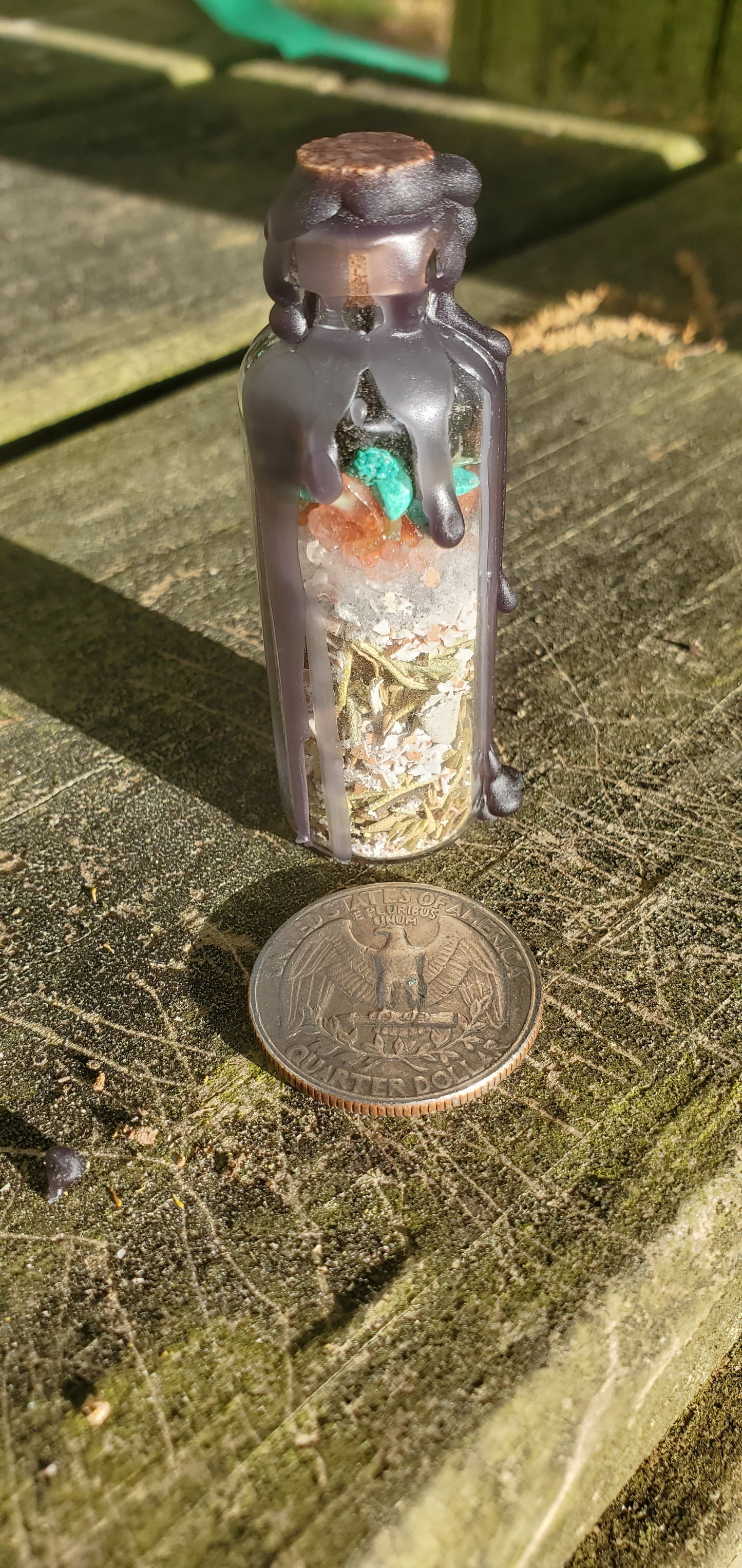 A glass bottle filled with herbs, crystals, and powders sealed with black wax. There is a quarter laying next to the bottle.