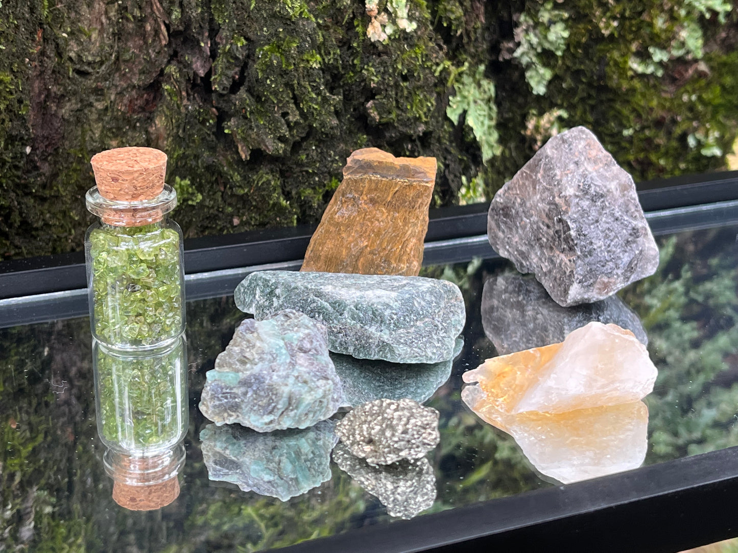 Raw emerald, raw pyrite, raw green aventurine, raw citrine, a bottle of tumbled peridot chips, raw tiger's eye, and raw smoky quartz set on a mirror with part of a tree as the backdrop.