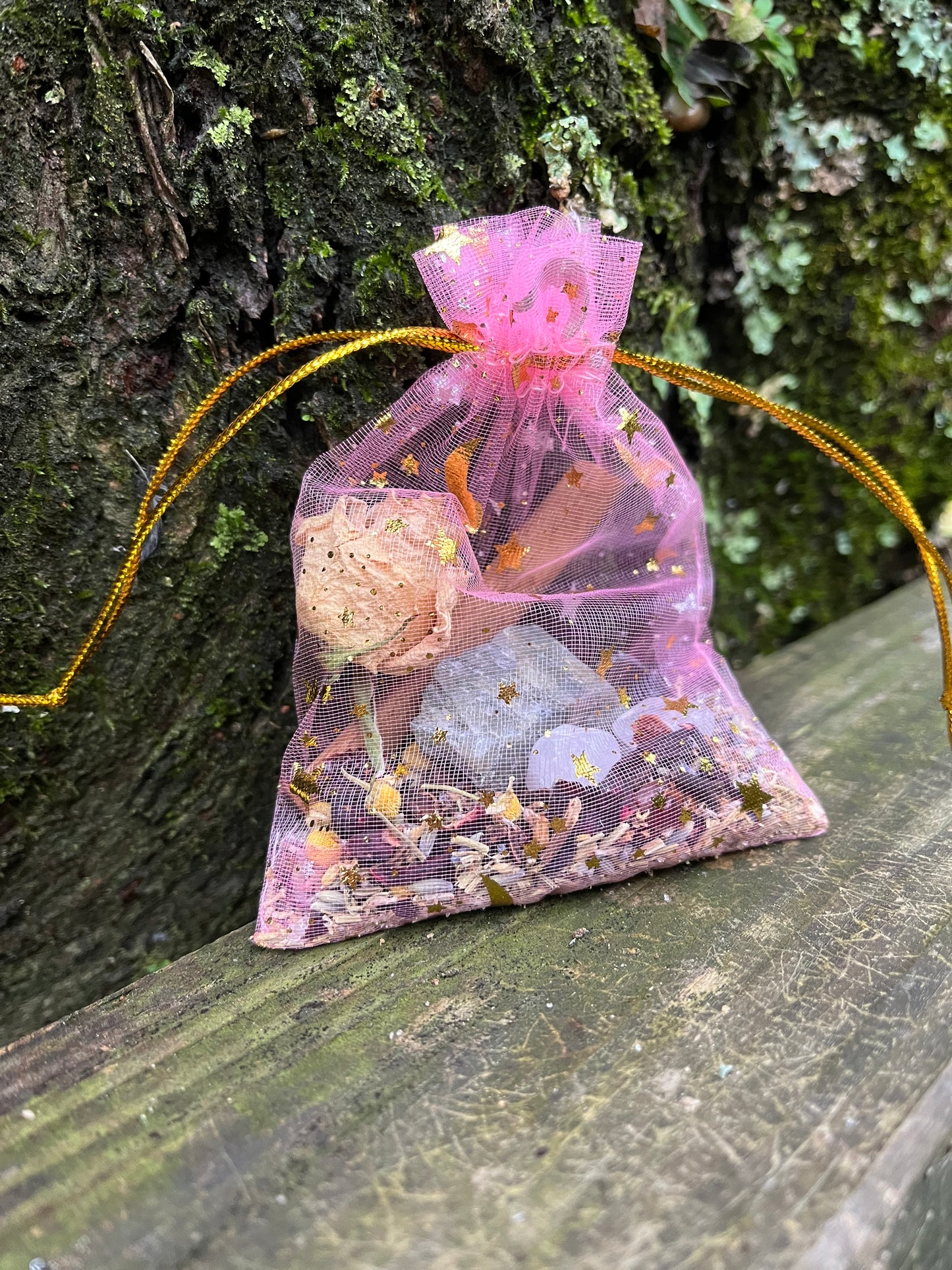 A pink organza bag with golden strings and gold stars and moons on it filled with herbs and crystals.
