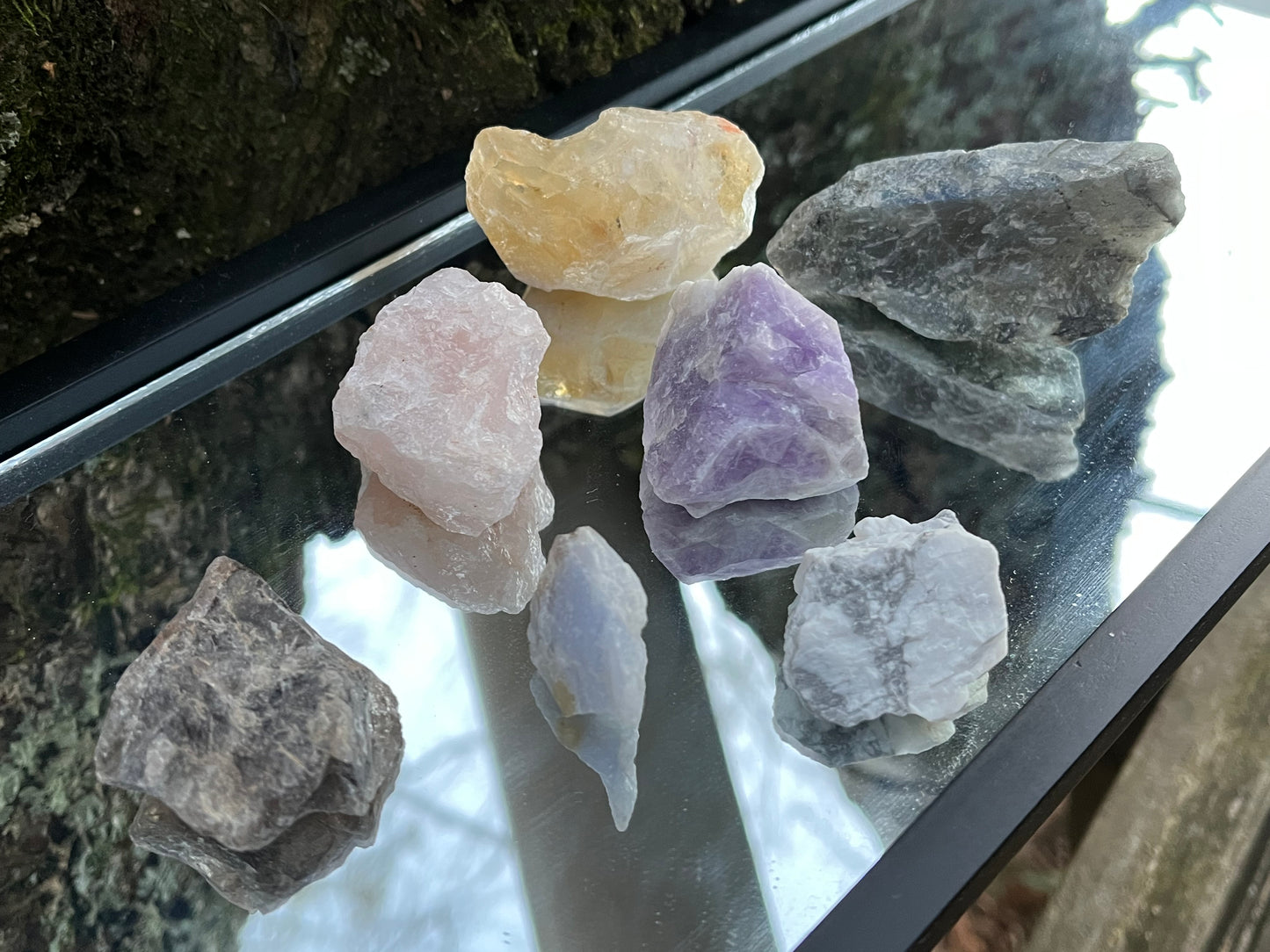 Raw amethyst, raw rose quartz, raw blue lace agate, raw citrine, raw smoky quartz, raw howlite, and raw labradorite set on a mirror with part of a tree as the backdrop.