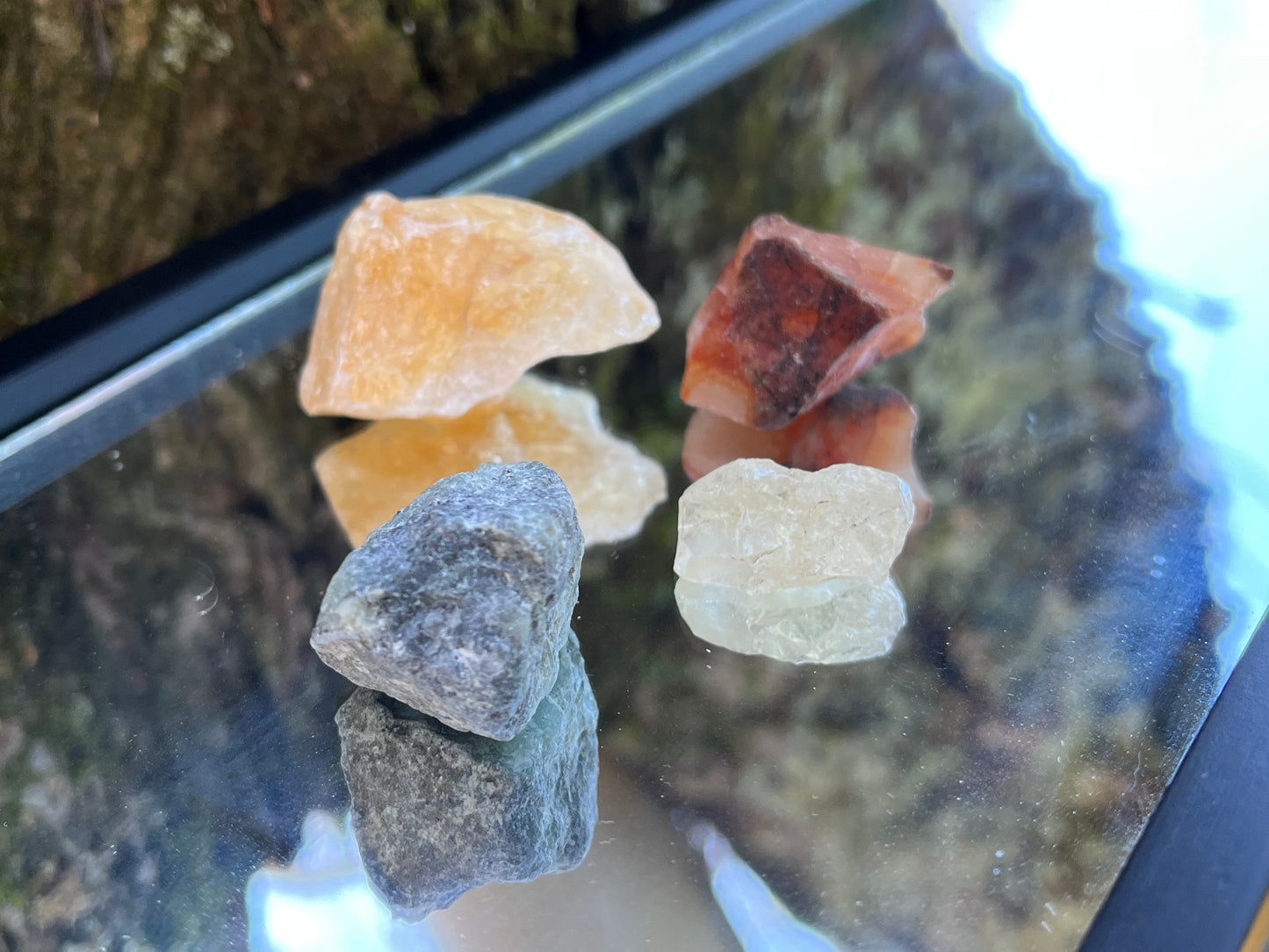 Raw citrine, raw carnelian, raw emerald, and raw clear quartz set on a mirror with part of a tree as the backdrop.