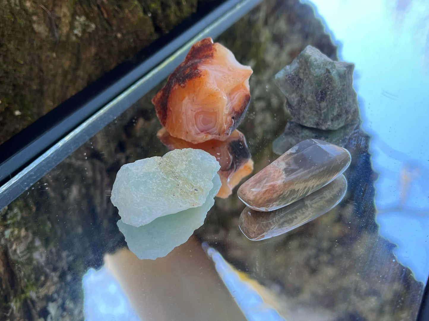 Raw carnelian, raw emerald, raw aquamarine, and tumbled black moonstone set on a mirror with part of a tree as the backdrop.