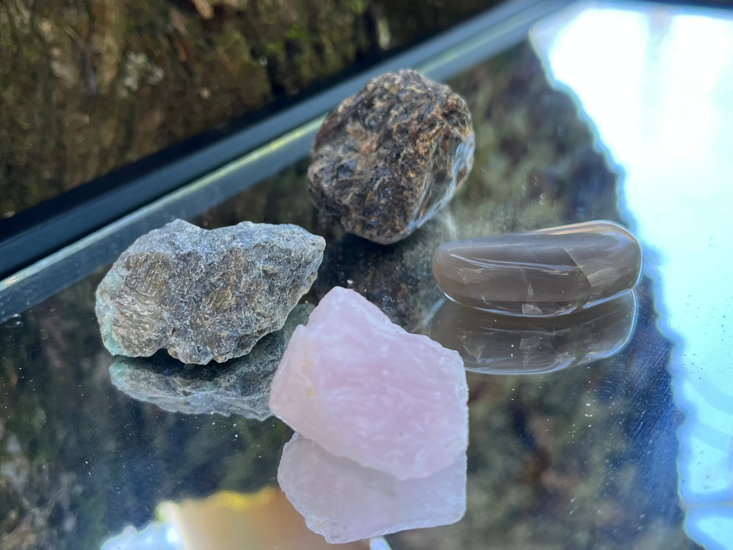 Raw black amber, raw emerald, raw rose quartz, and tumbled black moonstone set on a mirror with part of a tree as the backdrop.