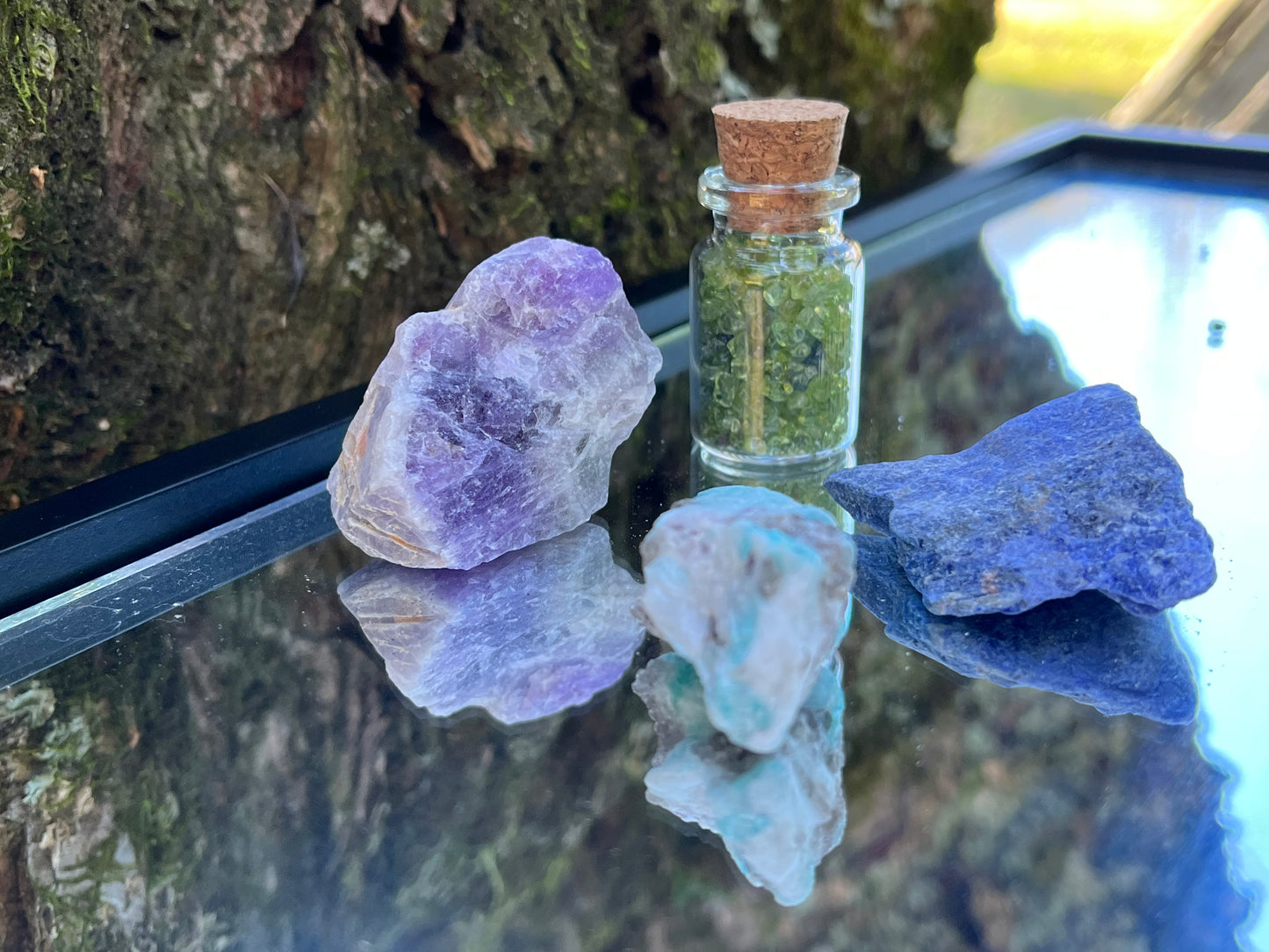 Raw amethyst, raw amazonite, raw sodalite, and a bottle of tumbled peridot chips set on a mirror with part of a tree as the backdrop.