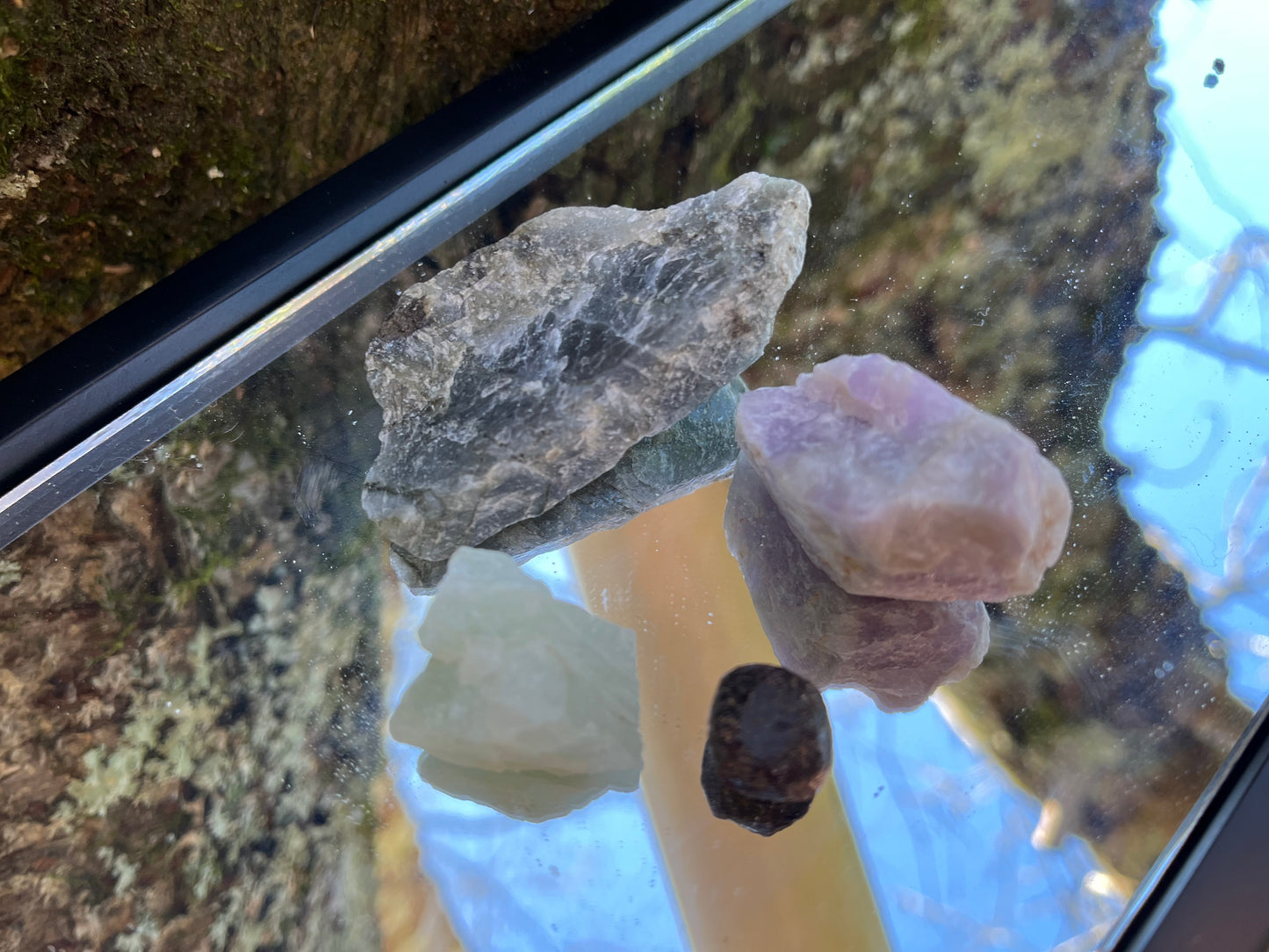 Raw labradorite, raw amethyst, raw aquamarine, and raw garnet set on a mirror with part of a tree as the backdrop.
