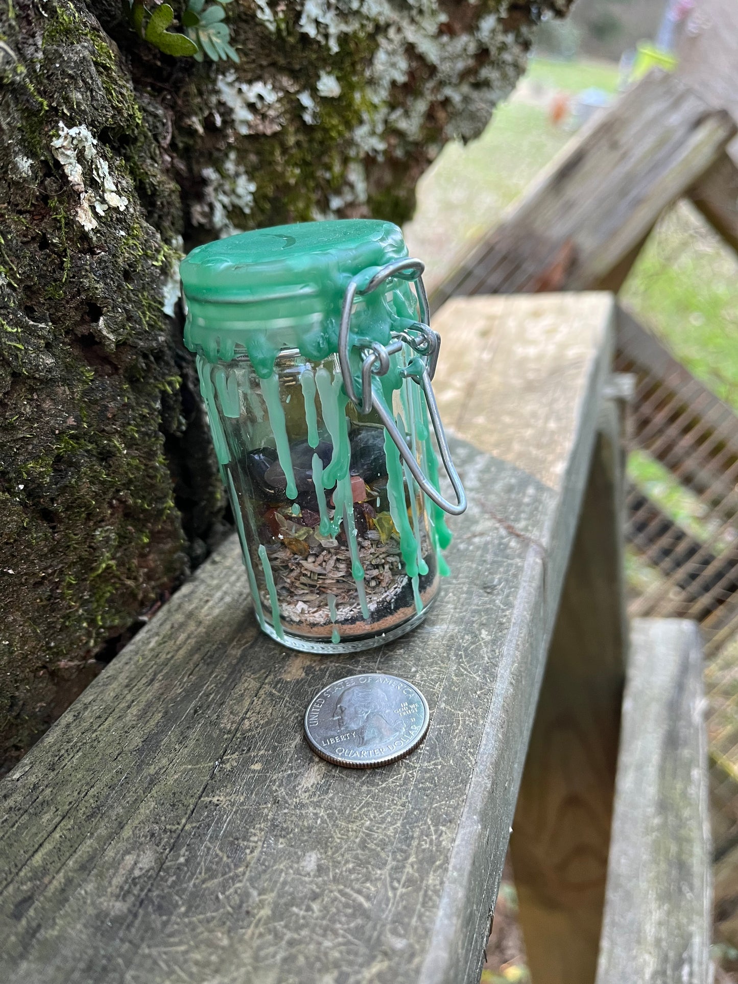 A tall glass jar with a clasp lid filled with herbs, crystals, and powders sealed with green wax. There is a quarter laying next to the jar.