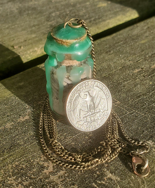 A glass bottle filled with herbs, crystals, and powders, sealed with green wax and on a chain. There is a quarter leaning against the bottle.