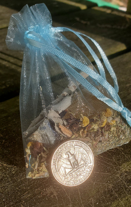 A blue organza bag with light blue strings filled with herbs and crystals. There is a quarter leaning against part of the bag.