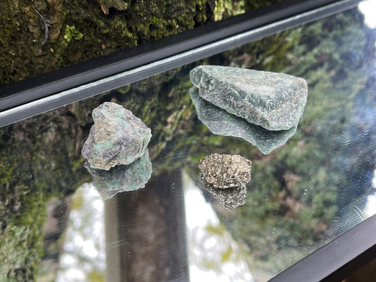 Raw emerald, raw pyrite, and raw green aventurine set on a mirror with part of a tree as the backdrop.