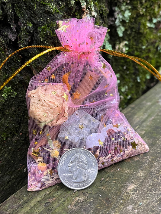 A pink organza bag with golden strings and gold stars and moons on it filled with herbs and crystals. There is a quarter leaning on part of the bag.