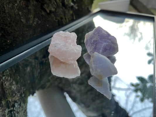 Raw amethyst, raw rose quartz, and raw blue lace agate set on a mirror with part of a tree as the backdrop.