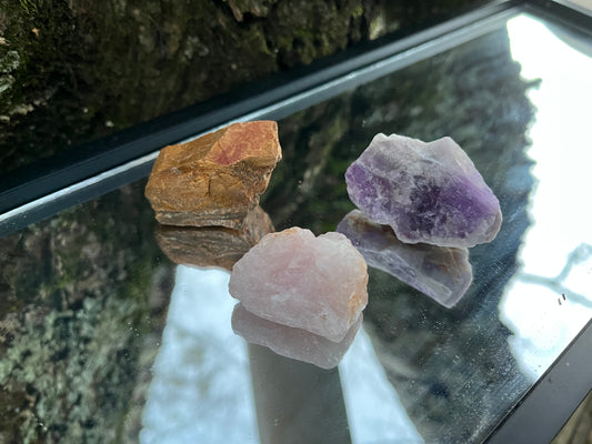 Raw tiger's eye, raw amethyst, and raw rose quartz set on a mirror with part of a tree as the backdrop.