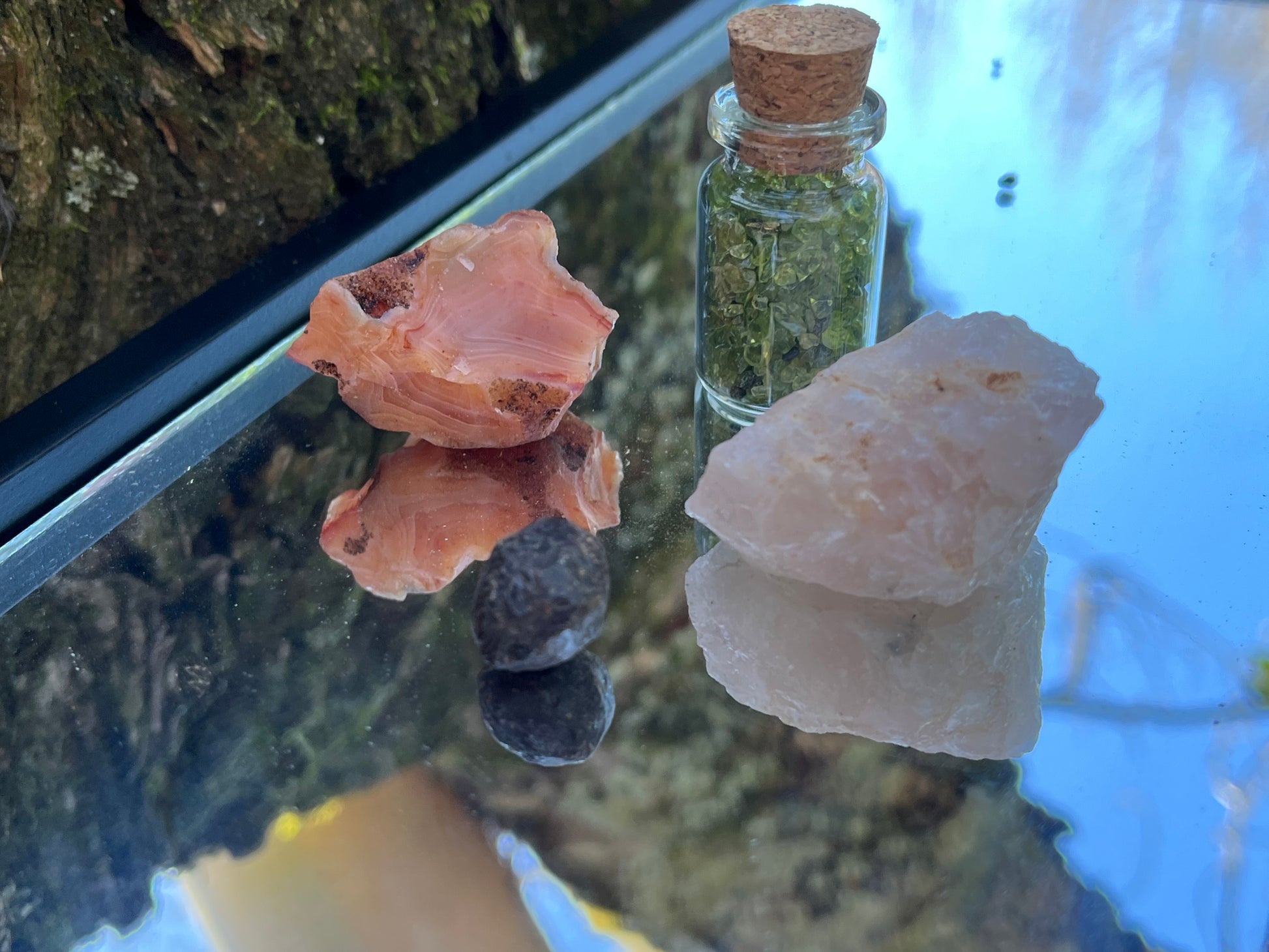 Raw carnelian, raw rose quartz, raw garnet, and a bottle of tumbled peridot chips set on a mirror with part of a tree as the backdrop.