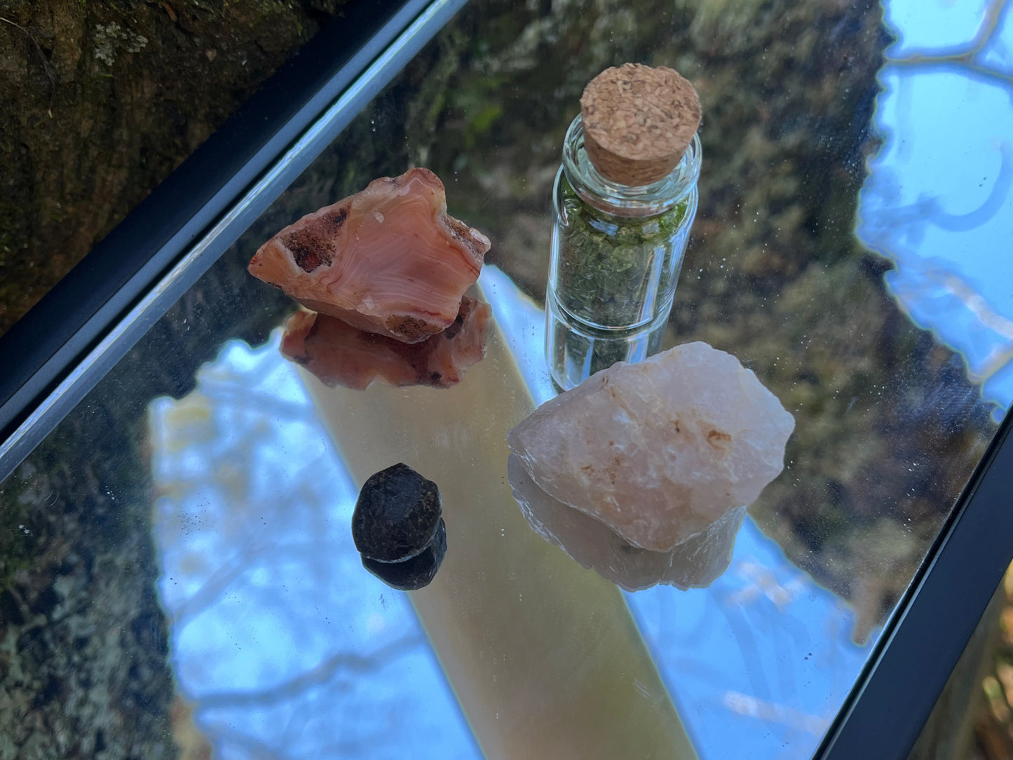 Raw carnelian, raw rose quartz, raw garnet, and a bottle of tumbled peridot chips set on a mirror with part of a tree as the backdrop.