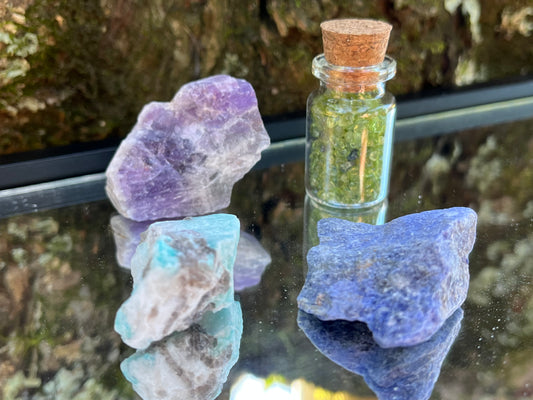 Raw amethyst, raw amazonite, raw sodalite, and a bottle of tumbled peridot chips set on a mirror with part of a tree as the backdrop.