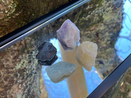 Raw black tourmaline, raw aquamarine, raw amethyst and raw citrine set on a mirror with part of a tree as the backdrop.