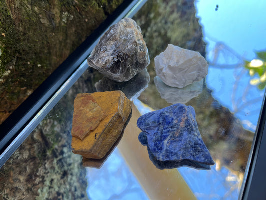 Raw smoky quartz, raw tiger's eye, raw howlite, and raw sodalite set on a mirror with part of a tree as the backdrop.