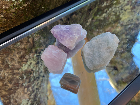 Raw amethyst, raw rose quartz, raw aquamarine, and tumbled black moonstone set on a mirror with part of a tree as the backdrop.