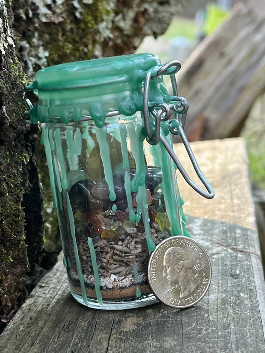 A tall glass jar with a clasp lid filled with herbs, crystals, and powders sealed with green wax. There is a quarter leaning against the jar.