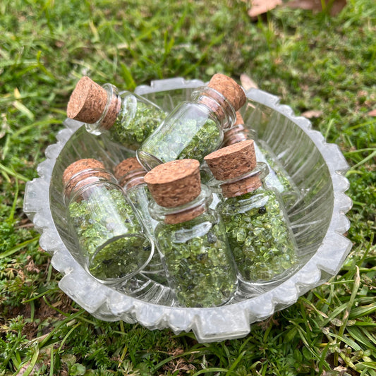 A small glass bowl full of tiny glass bottles with cork stoppers full of green peridot chips.