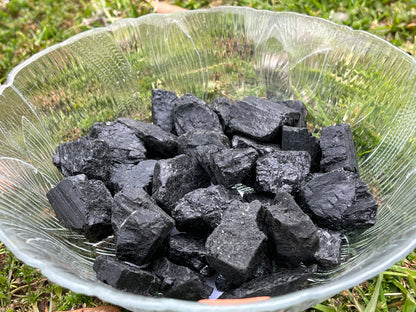 Several pieces of mini raw black tourmaline in a round glass bowl. The bowl is set on grass.