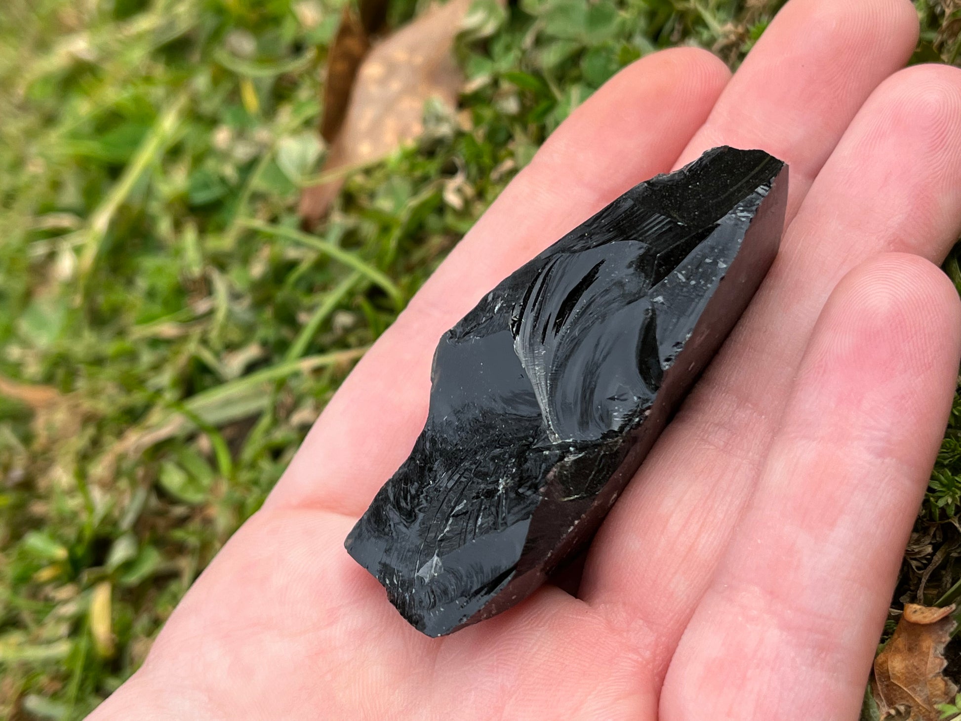 A piece of raw black obsidian on a sand to show size. It is about 1 inch wide and 2.5 inches long.