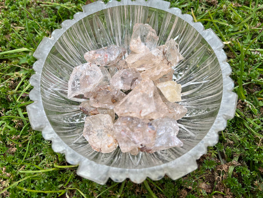 Small pieces of raw clear quartz in a small, round, glass bowl. The bowl is set in grass.