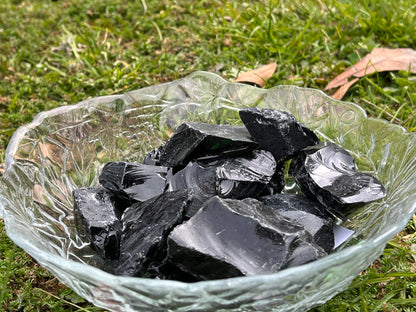 Pieces of raw black obsidian in a round glass bowl. The bowl is sitting in some grass.