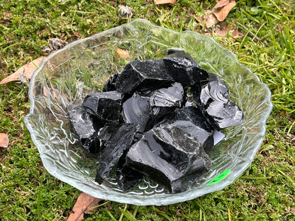 Pieces of raw black obsidian in a round glass bowl. The bowl is sitting in some grass.