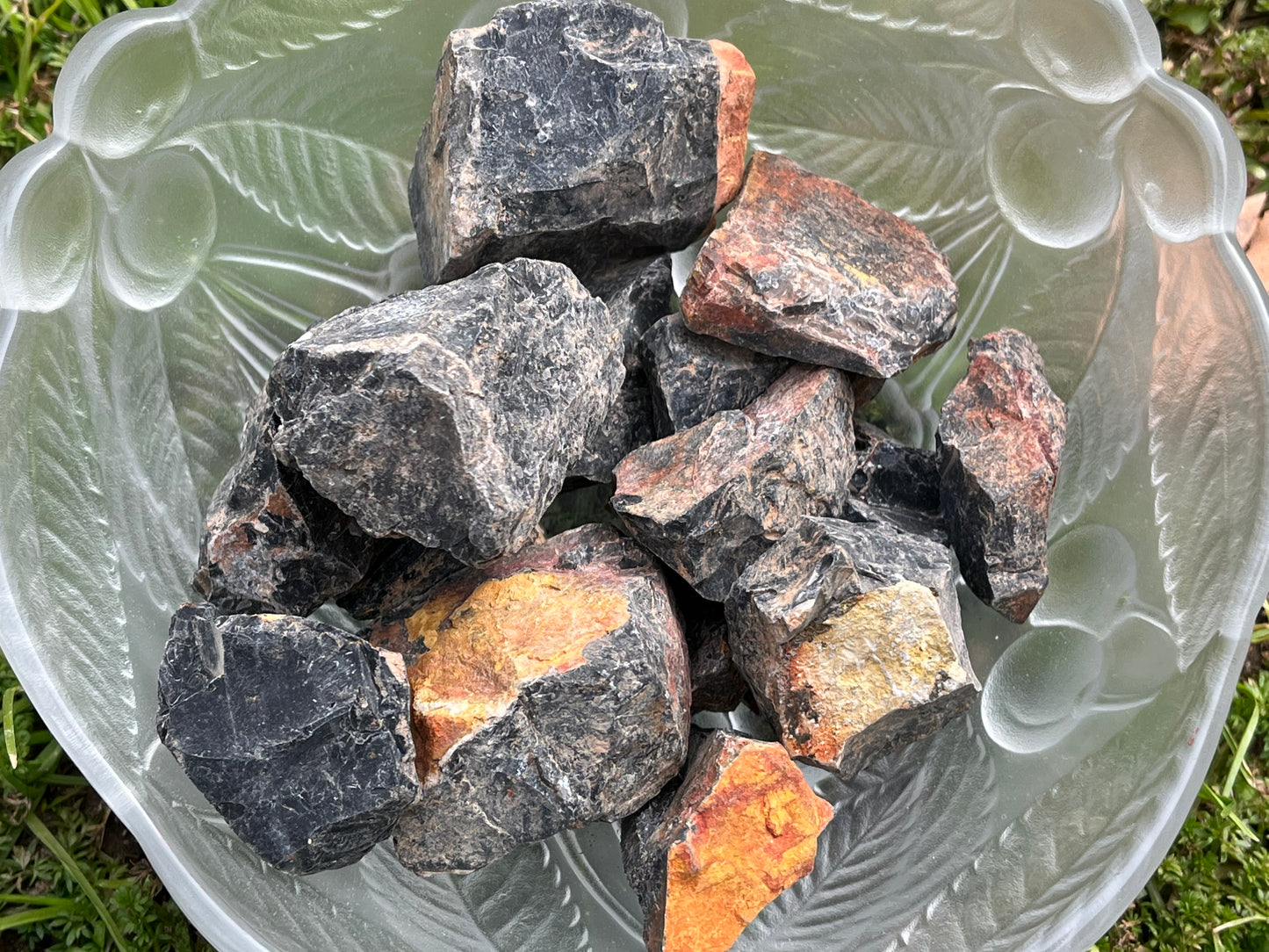 Pieces of raw black obsidian in a round glass bowl. The bowl is sitting in some grass.